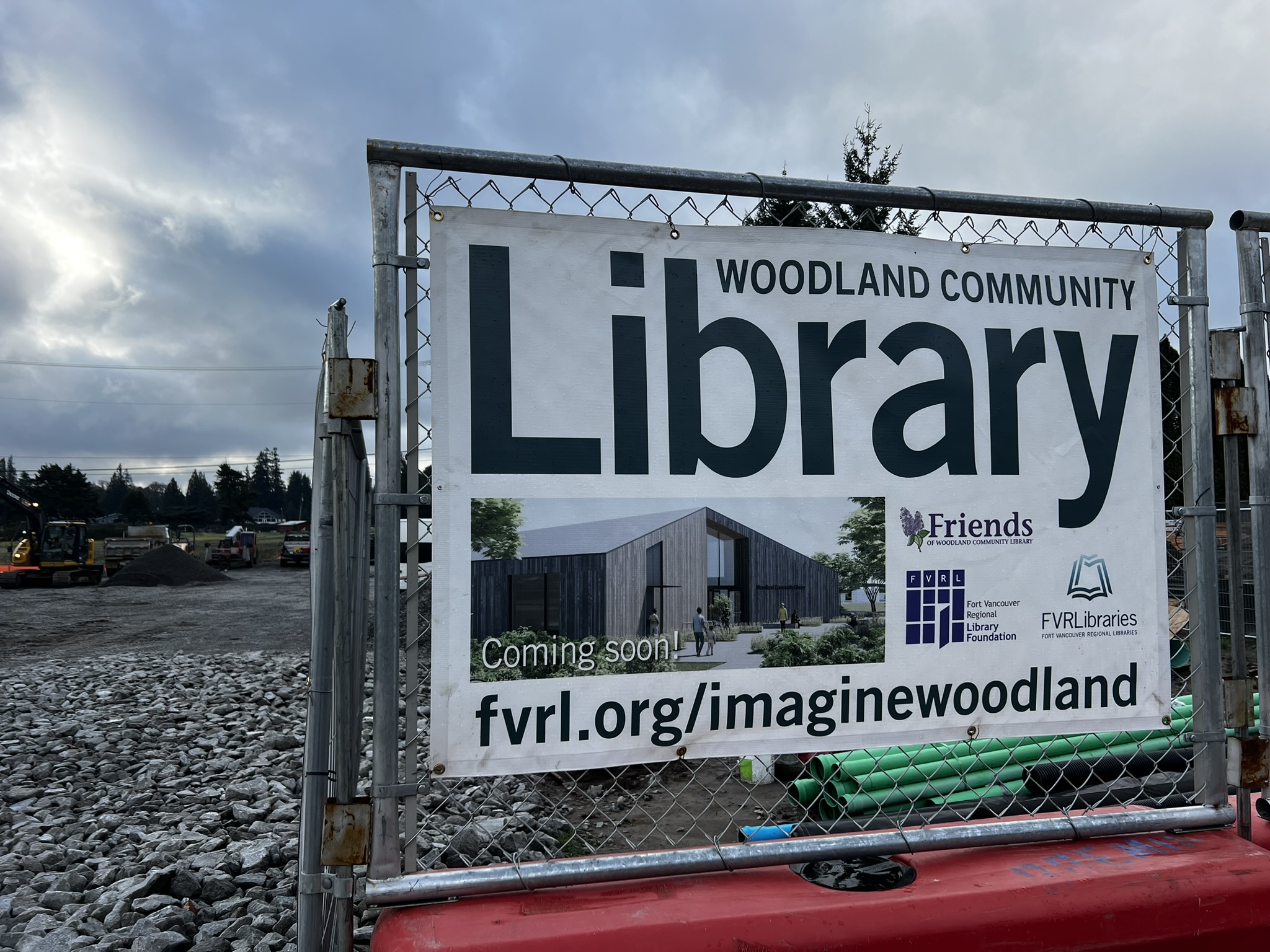 Sign posted to a chain link fence. The sign reads: "Woodland Community Library—coming soon!" 