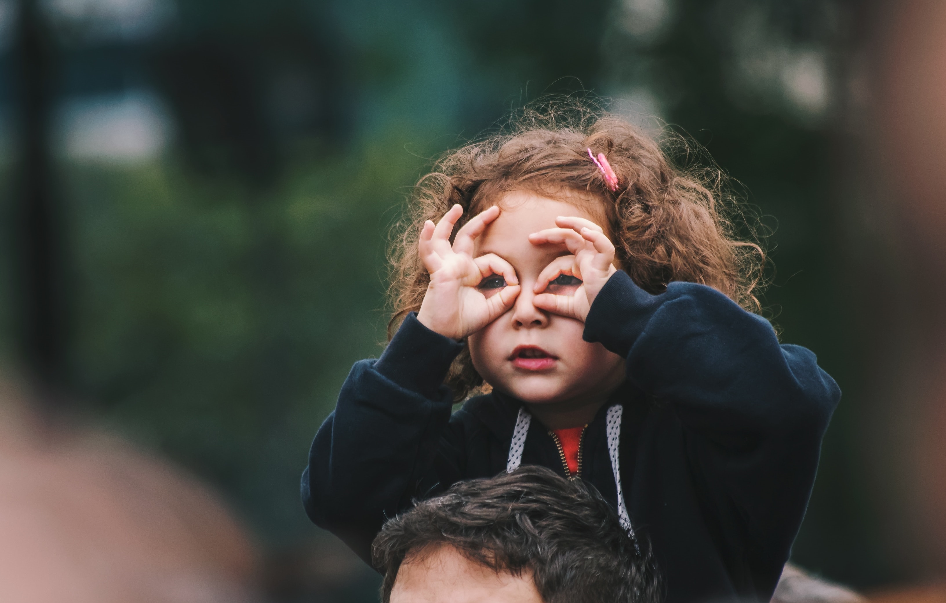 Picture of a child perched on an adult's shoulders, circling her thumbs  and index fingers around her eyes to mimic glasses. 