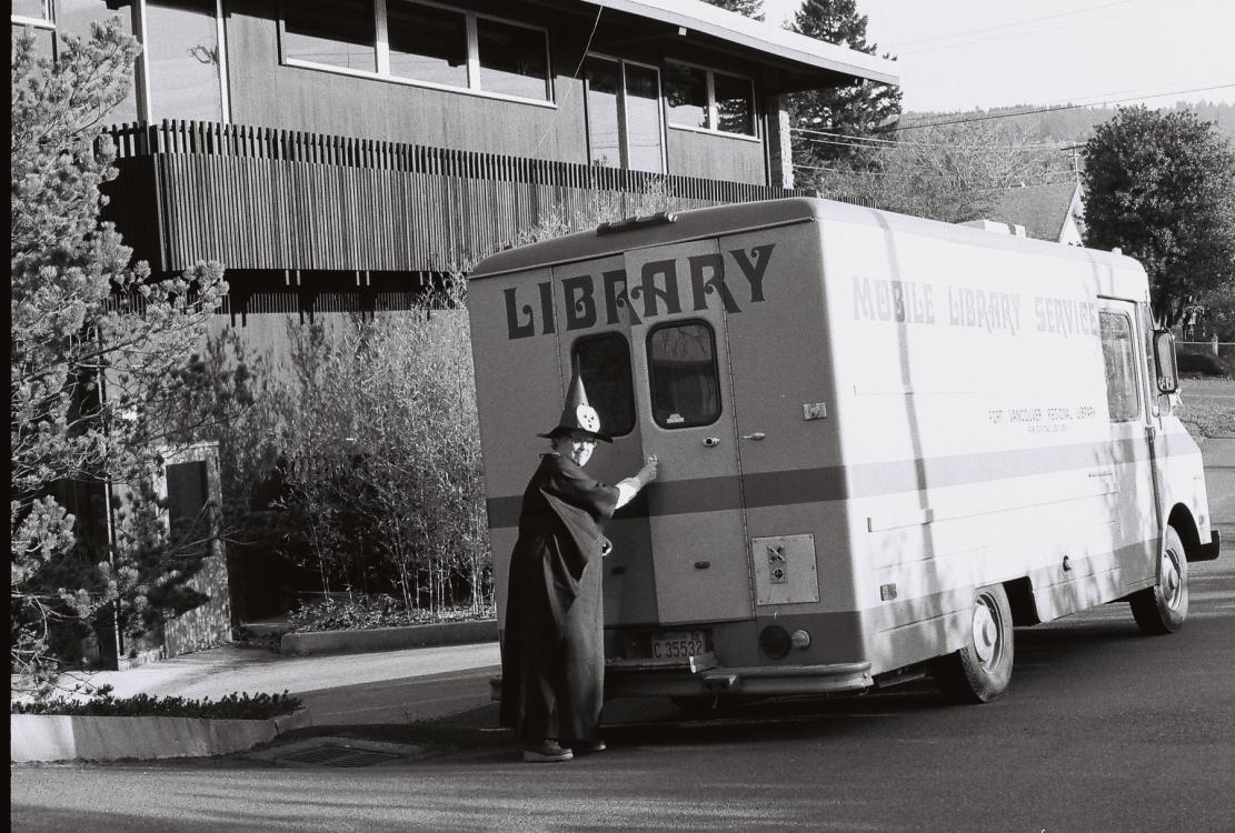 Black-and-white photo of a worker opening the rear doors of a FVRLibraries bookmobile, circa 1973. The worker is wearing a witch costume, including a pointy black hat with a paper Jack-o'-lantern attached to it.